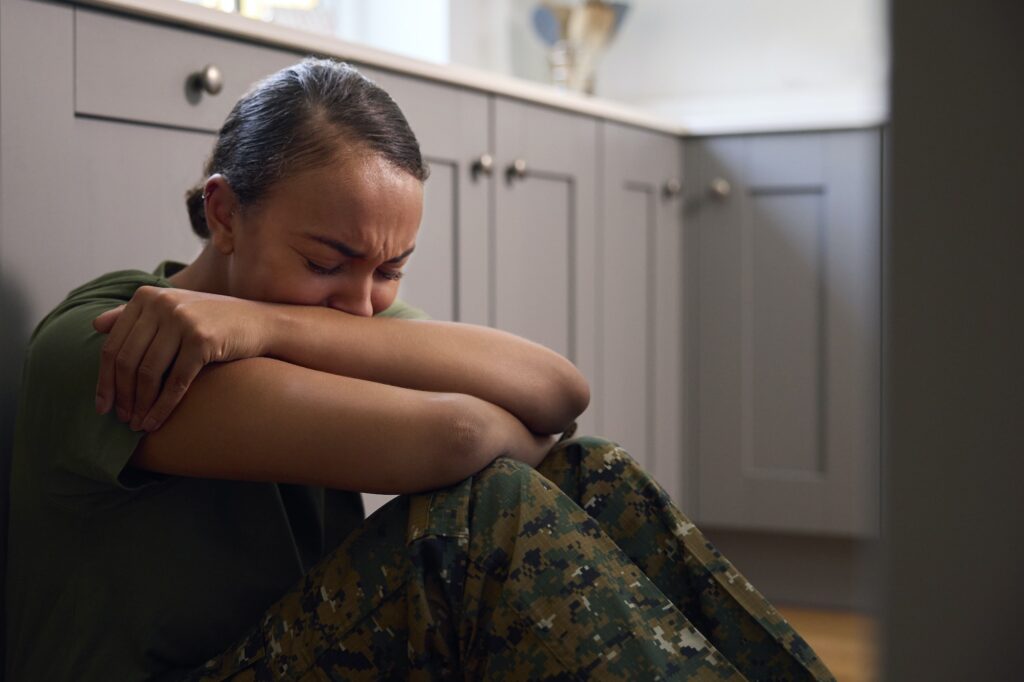 Depressed Female Soldier In Uniform Suffering With PTSD Sitting On Kitchen Floor On Home Leave